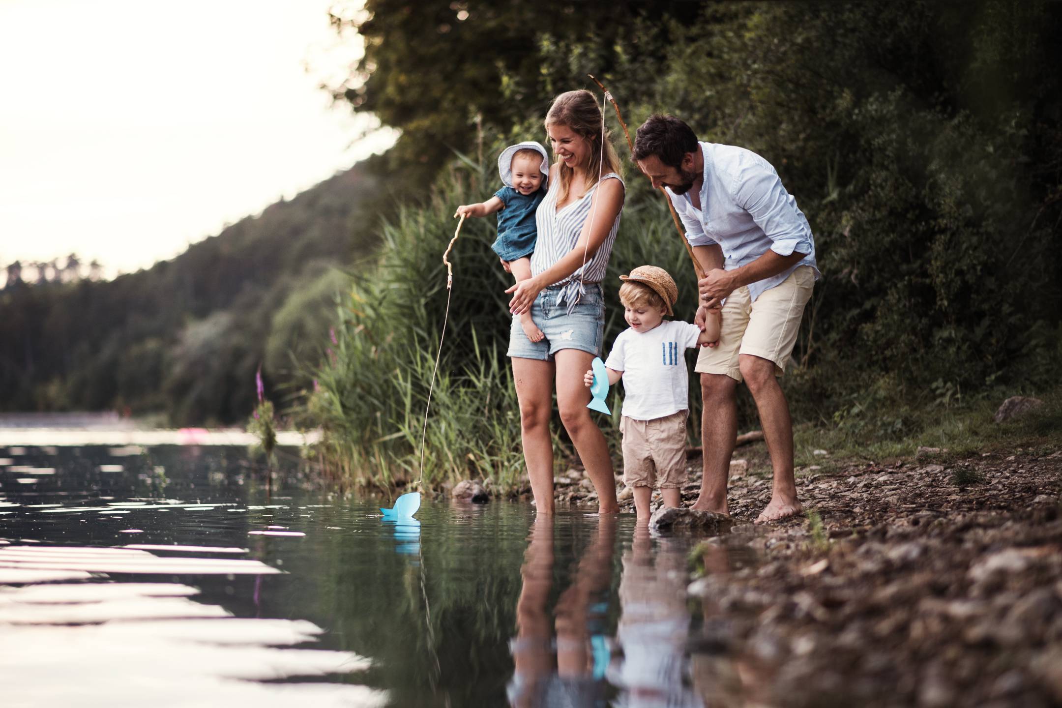 A young couple with two young children standing by a river while holding sticks made to look like fishing poles.