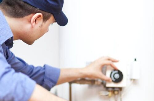 A plumber crouching next to a residential water heater and using one hand to turn a dial on the heater.