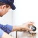 A plumber crouching next to a residential water heater and using one hand to turn a dial on the heater.