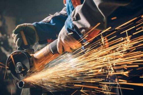 A person wearing gloves and other protective gear works with a metalwork grinder. Sparks are flying around them.