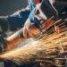 A person wearing gloves and other protective gear works with a metalwork grinder. Sparks are flying around them.