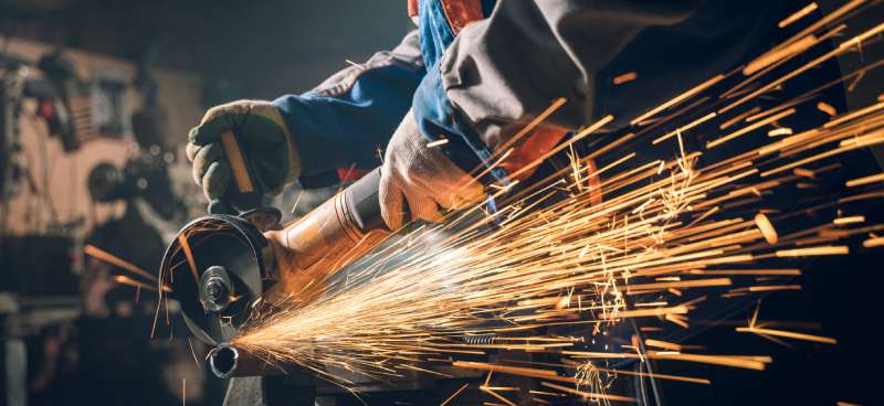 A person wearing gloves and other protective gear works with a metalwork grinder. Sparks are flying around them.