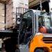 A worker is driving an orange and grey forklift. They're using it to remove a pallet of boxes from a truck.