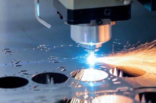 A laser cutter works hard, cutting multiple circles from a piece of thick, dark-colored metal. Sparks are flying from the laser.