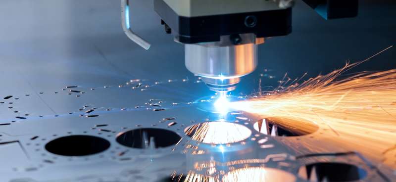 A laser cutter works hard, cutting multiple circles from a piece of thick, dark-colored metal. Sparks are flying from the laser.