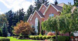 Roof and Home Security The exterior of a red brick house with beautifully kept landscaping including well-shaped bushes, trees, and potted flowers.