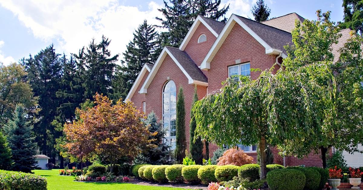The exterior of a red brick house with beautifully kept landscaping including well-shaped bushes, trees, and potted flowers.