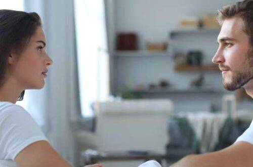 A young man and woman, both in white T-shirts, sitting across from one another in a living room as they look at one another.