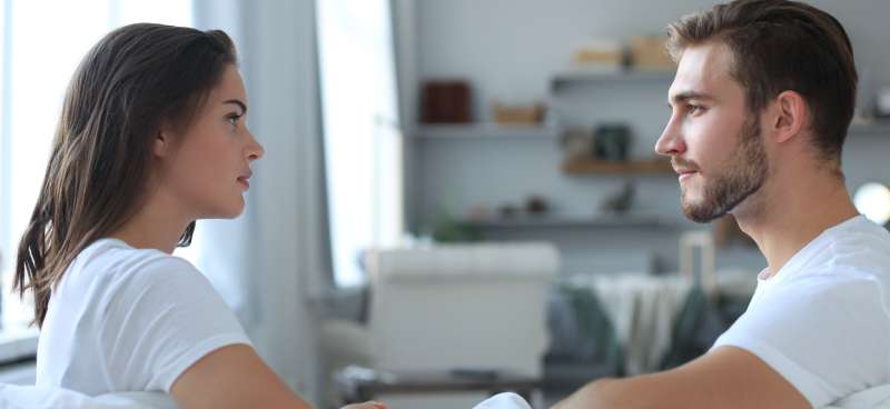 A young man and woman, both in white T-shirts, sitting across from one another in a living room as they look at one another.