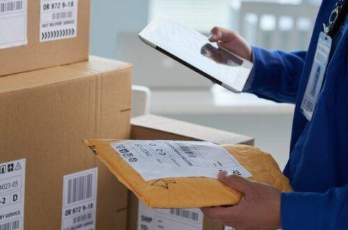 A worker in a blue uniform stands next to a stack of cardboard boxes with shipping labels. The worker is holding a tablet.