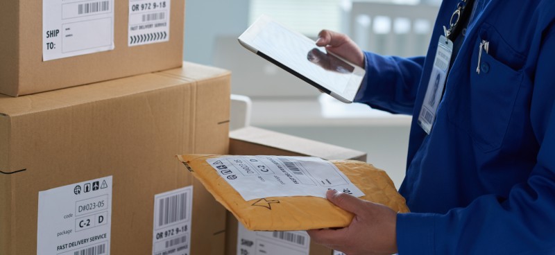 A worker in a blue uniform stands next to a stack of cardboard boxes with shipping labels. The worker is holding a tablet.