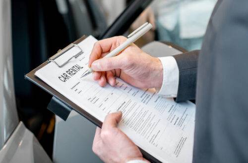 A man wearing a suit holds a clipboard with a contract reading "car rental" in a showroom next to a silver sedan.