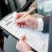 A man wearing a suit holds a clipboard with a contract reading "car rental" in a showroom next to a silver sedan.