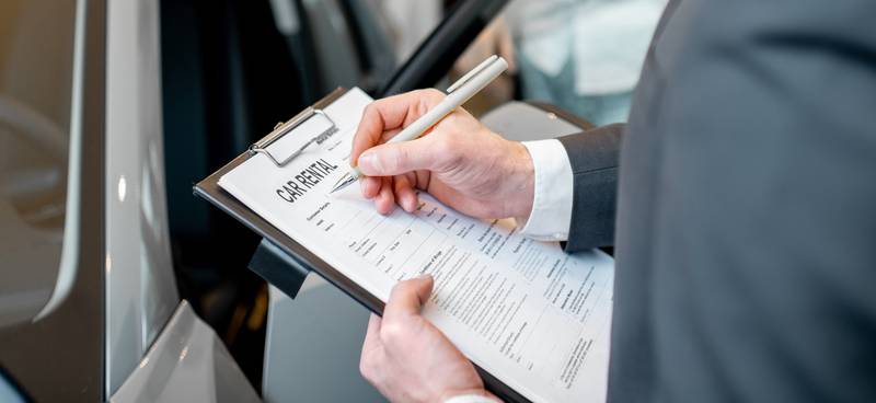 A man wearing a suit holds a clipboard with a contract reading "car rental" in a showroom next to a silver sedan.