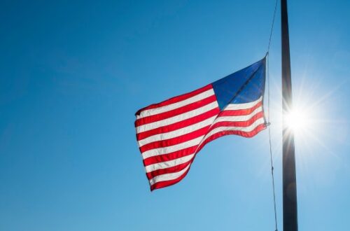 An American flag flying on a flagpole against a clear blue sky. The flag is lowered to half-mast on the pole.