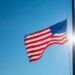 An American flag flying on a flagpole against a clear blue sky. The flag is lowered to half-mast on the pole.