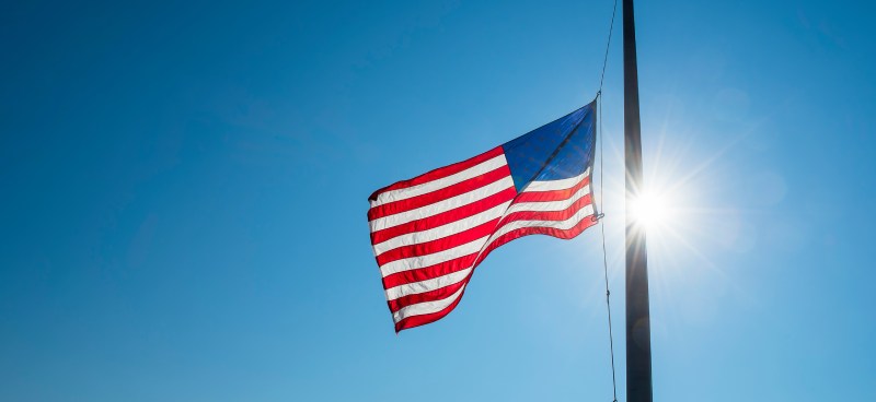 An American flag flying on a flagpole against a clear blue sky. The flag is lowered to half-mast on the pole.