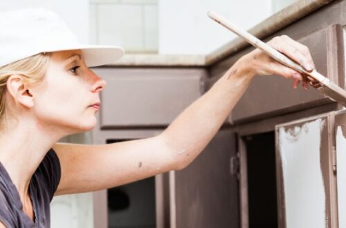 A woman wearing a white baseball cap holds a paint brush to paint her lower kitchen cabinets chocolate brown.