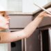 A woman wearing a white baseball cap holds a paint brush to paint her lower kitchen cabinets chocolate brown.