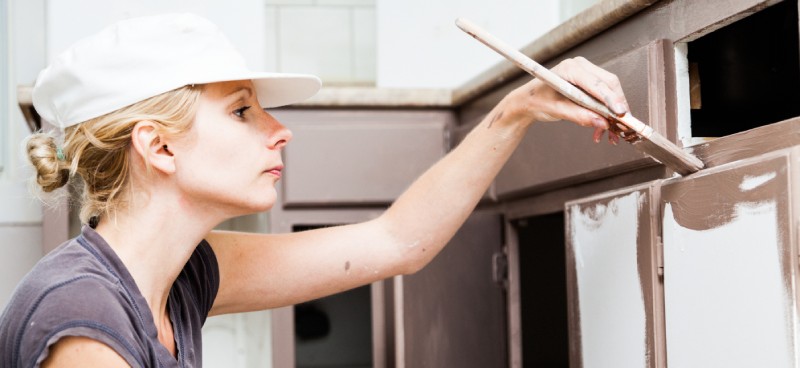 A woman wearing a white baseball cap holds a paint brush to paint her lower kitchen cabinets chocolate brown.