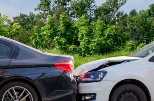 A white sedan colliding with the rear end of a black sedan with grass, trees, and a cloudy sky in the background.