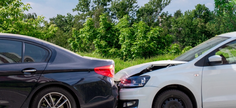 A white sedan colliding with the rear end of a black sedan with grass, trees, and a cloudy sky in the background.