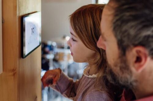 A father and a daughter look at their smart home thermostat displaying "19 degrees" against a wooden beam in their home.