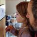 A father and a daughter look at their smart home thermostat displaying "19 degrees" against a wooden beam in their home.