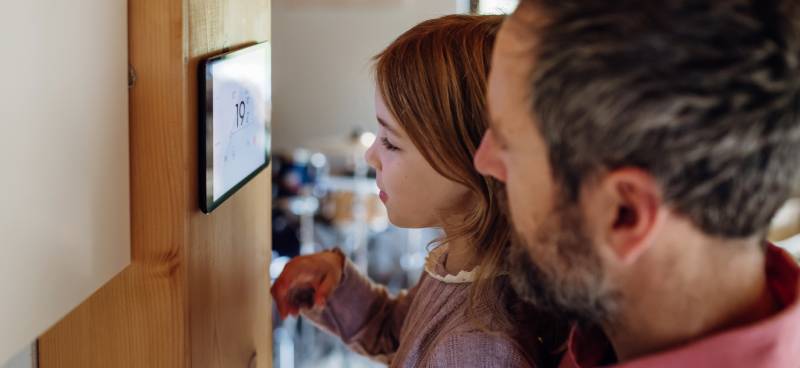 A father and a daughter look at their smart home thermostat displaying "19 degrees" against a wooden beam in their home.