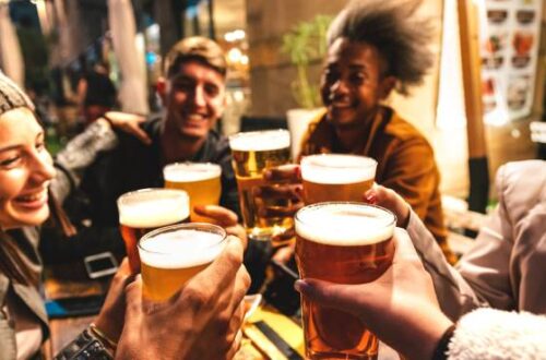 A group of four friends holding up their beers, celebrating while sitting in the outdoor area of a brewery.