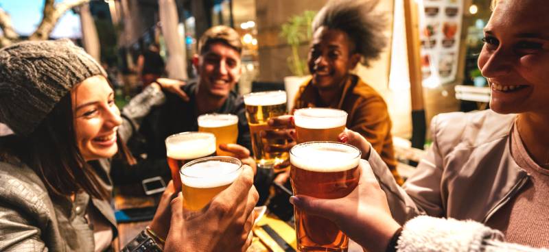 A group of four friends holding up their beers, celebrating while sitting in the outdoor area of a brewery.