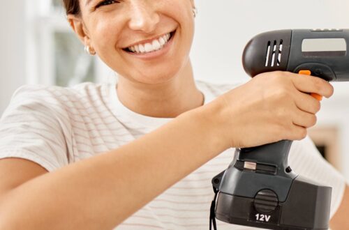 A woman is wearing a cream-colored striped t-shirt and smiling as she holds a power drill up to a wall.