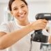 A woman is wearing a cream-colored striped t-shirt and smiling as she holds a power drill up to a wall.