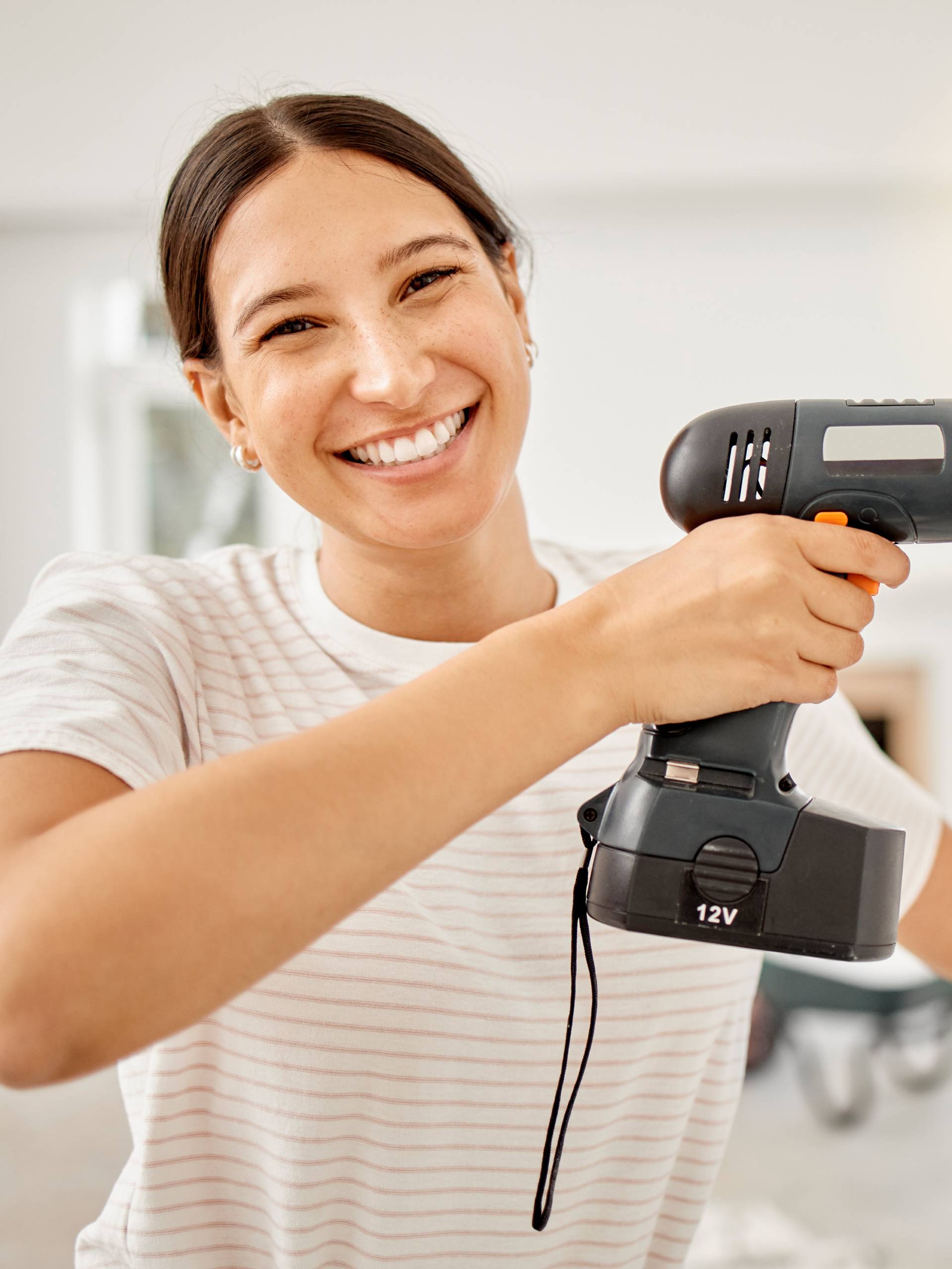 A woman is wearing a cream-colored striped t-shirt and smiling as she holds a power drill up to a wall.