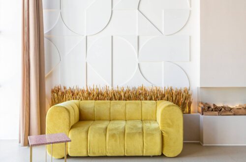 A modern loft apartment with large floor-to-ceiling windows, a yellow couch, and eccentric wall decor to the right.