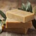 A stack of three homemade soap bars sitting on a wooden table. There's an olive branch and a few olives next to the stack.