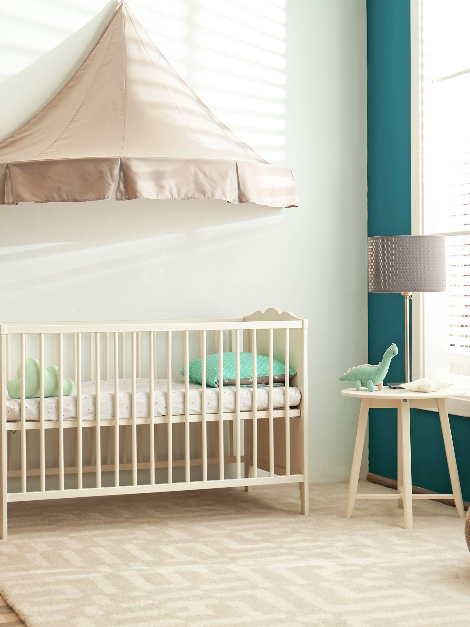 A neutral-themed baby nursery with a crib, a papillon chair with a yellow cushion, and large windows with blinds.