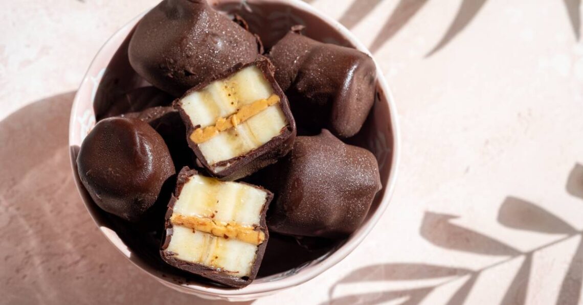 A bowl of dark chocolate-covered piece of banana on a table with a light shining through to create shadows of plants.