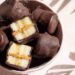 A bowl of dark chocolate-covered piece of banana on a table with a light shining through to create shadows of plants.