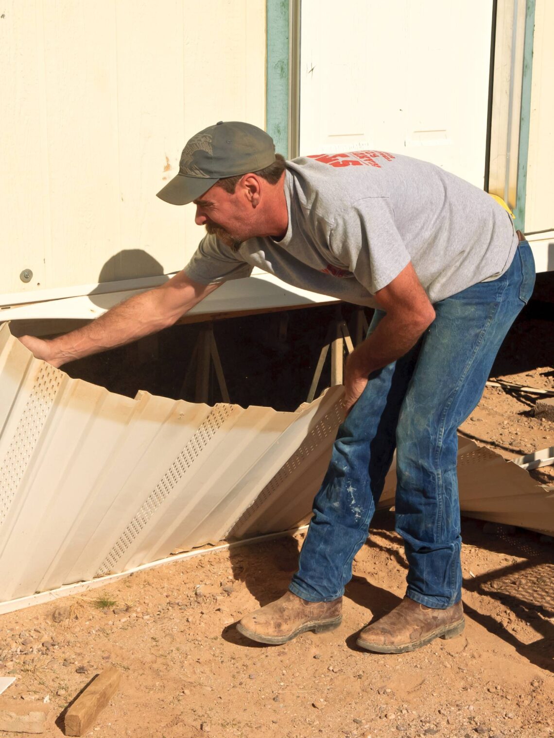 A man removes the skirting from an older mobile home during a renovation project. A black utility cart sits nearby.