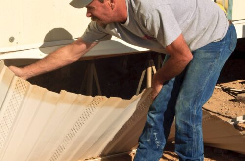 A man removes the skirting from an older mobile home during a renovation project. A black utility cart sits nearby.