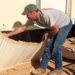 A man removes the skirting from an older mobile home during a renovation project. A black utility cart sits nearby.