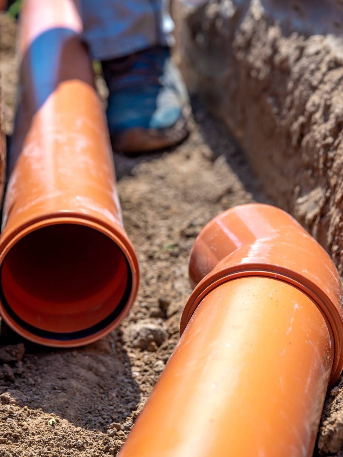 Two large orange drainage pipes lay in an underground excavated tunnel. A person's boot walks in between the area.