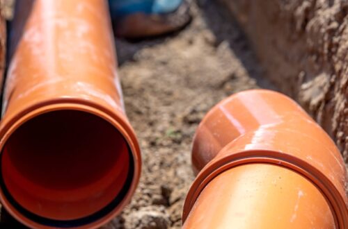 Two large orange drainage pipes lay in an underground excavated tunnel. A person's boot walks in between the area.