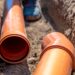 Two large orange drainage pipes lay in an underground excavated tunnel. A person's boot walks in between the area.