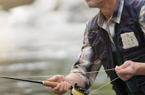 A man in waders, a plaid shirt, and a fishing vest fly fishing in a river. He is holding a fishing rod and pulling on the line.