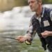 A man in waders, a plaid shirt, and a fishing vest fly fishing in a river. He is holding a fishing rod and pulling on the line.