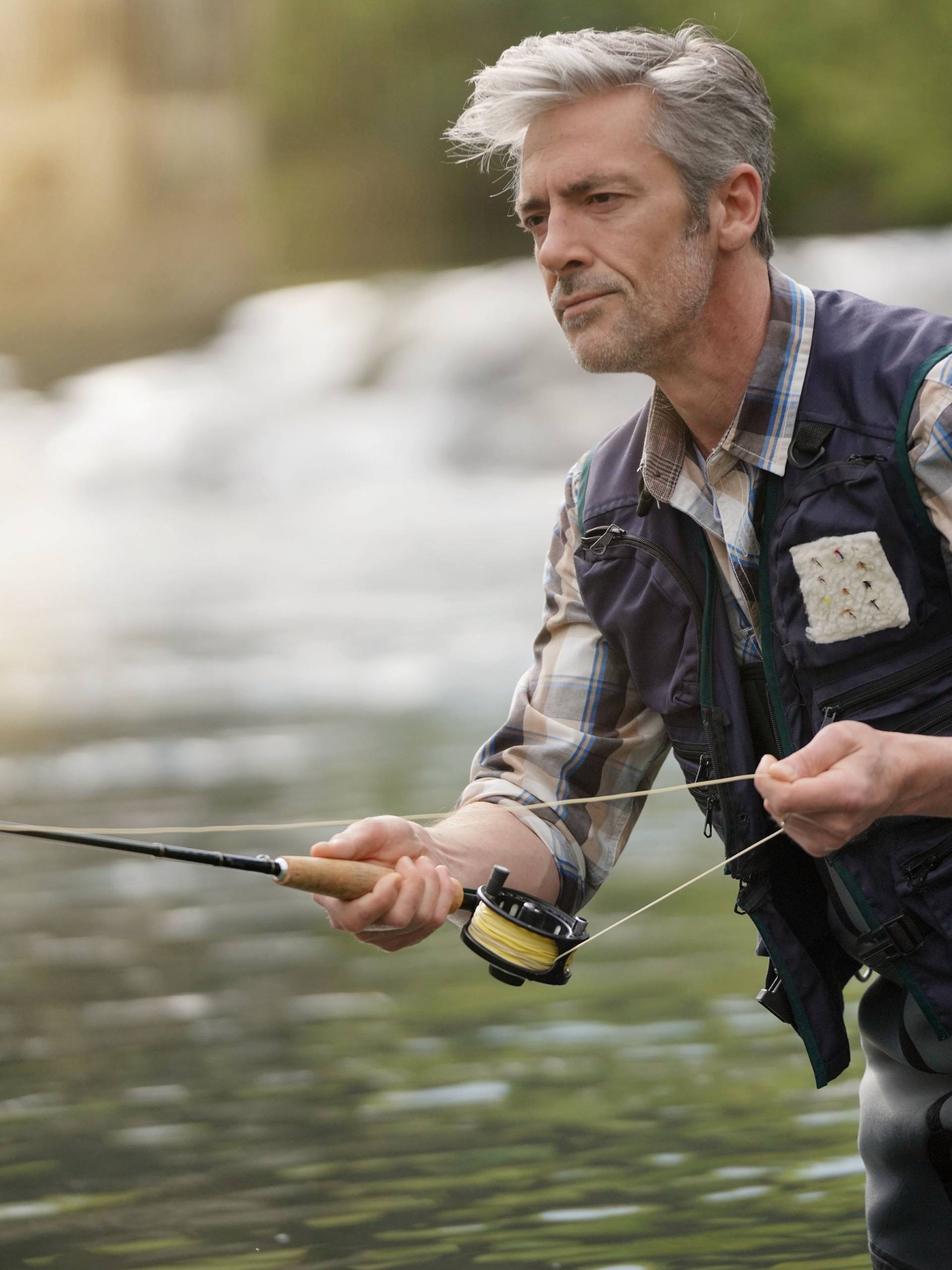 A man in waders, a plaid shirt, and a fishing vest fly fishing in a river. He is holding a fishing rod and pulling on the line.