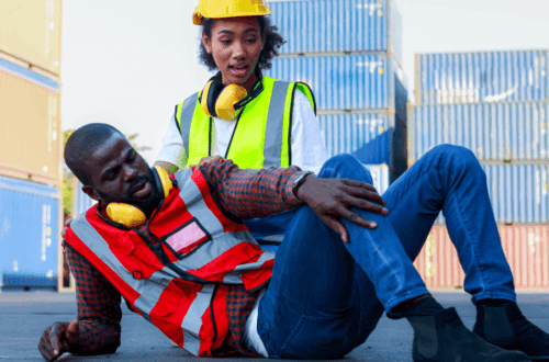 A Black male engineer is injured and on the ground, while a Black female engineer looks at his injury on his shin.