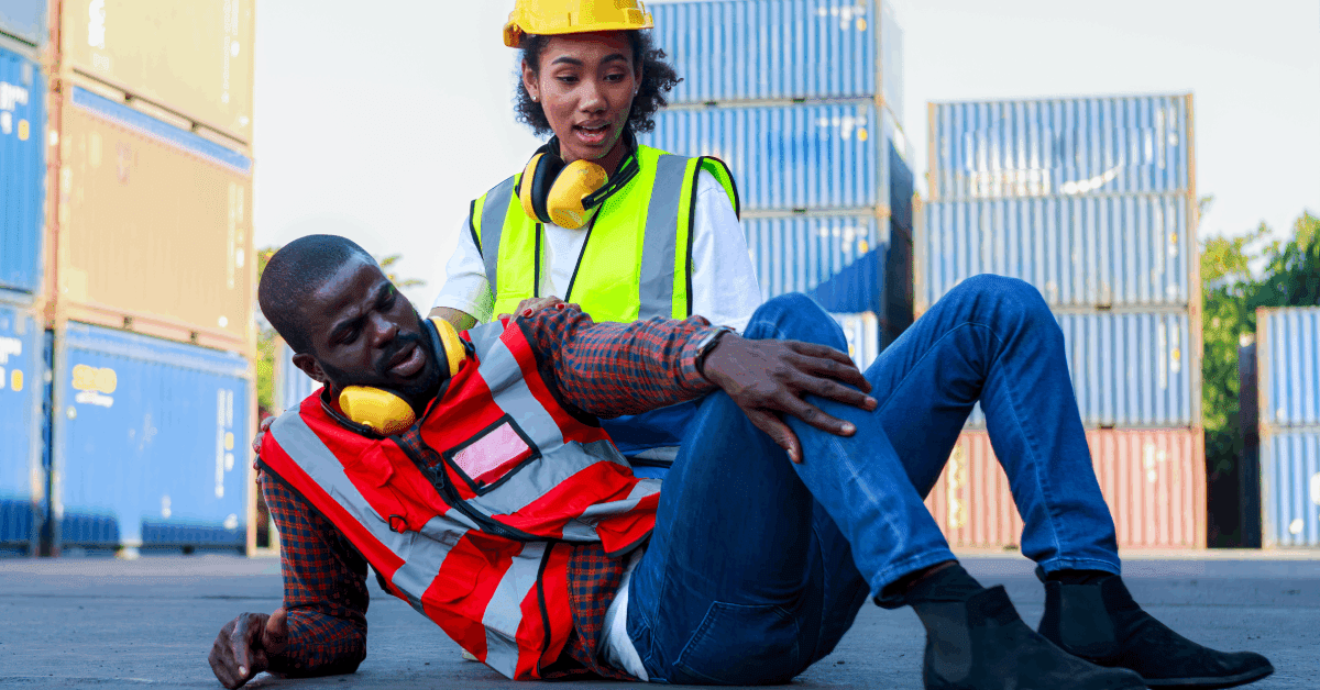 A Black male engineer is injured and on the ground, while a Black female engineer looks at his injury on his shin.